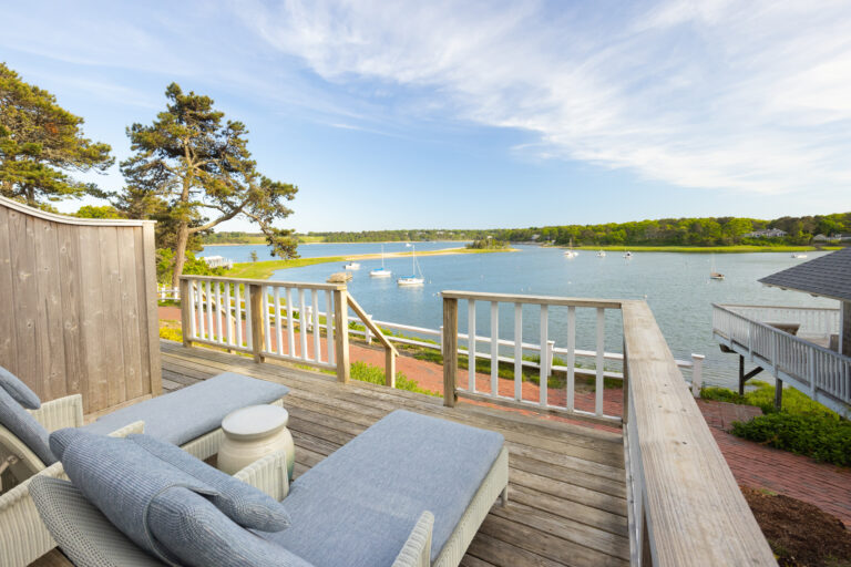 Wooden deck with lounge chairs overlooks a calm bay with sailboats, offering a stunning water view and easy access from the Queen Suite, bordered by trees and houses under a partly cloudy sky.