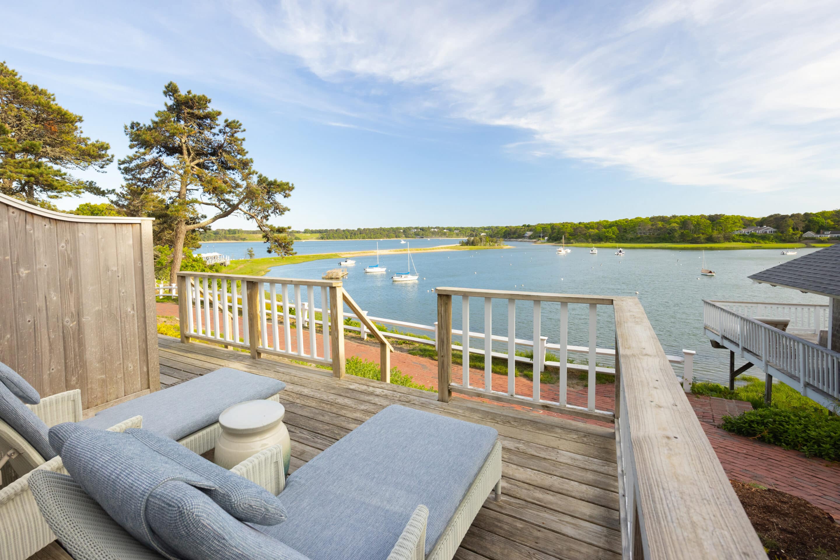 Wooden deck with lounge chairs overlooks a calm bay with sailboats, offering a stunning water view and easy access from the Queen Suite, bordered by trees and houses under a partly cloudy sky.