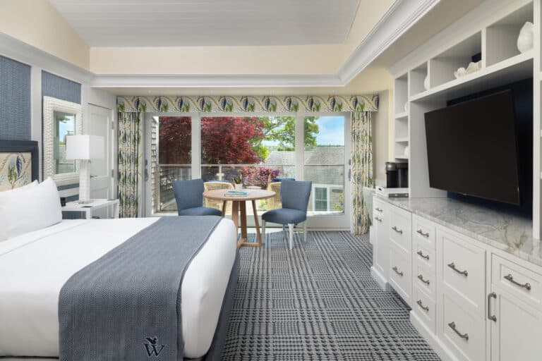 Hotel room with two queen beds, blue and white décor, a TV, a small round table with two chairs, and large windows overlooking a tree and rooftops, creating a relaxing Sweet Pea-inspired retreat.