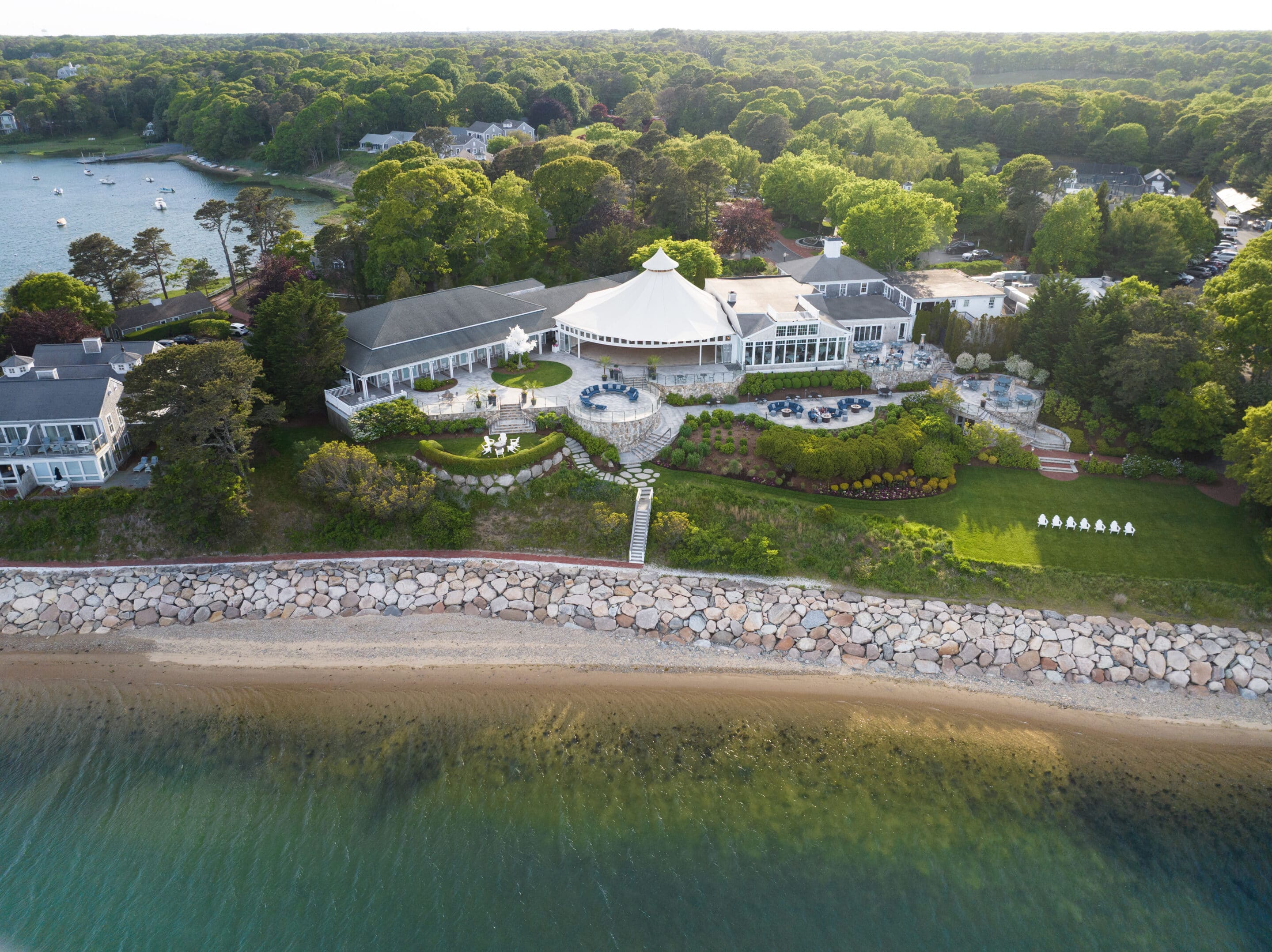 Aerial view of a large waterfront estate reminiscent of a five star resort, with multiple buildings, landscaped gardens, stone seawall, sandy beach, and tree-lined background along Cape Cod.
