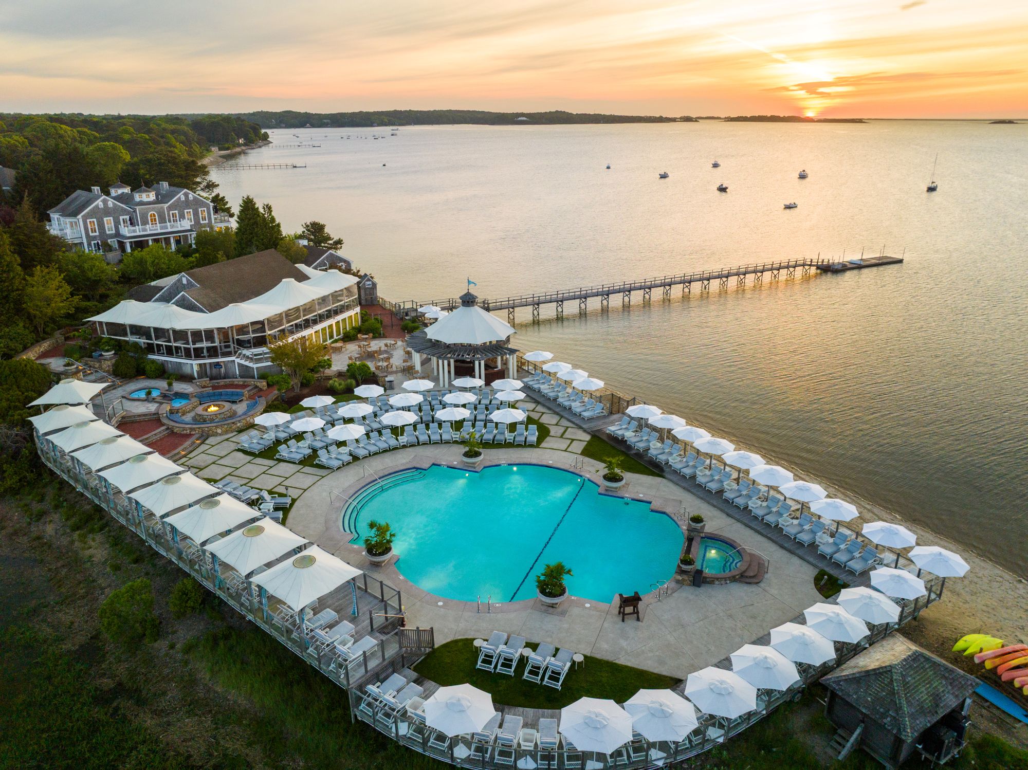 Aerial view of a large circular pool with white lounge chairs and umbrellas, near a lake with a dock and boats at sunset.