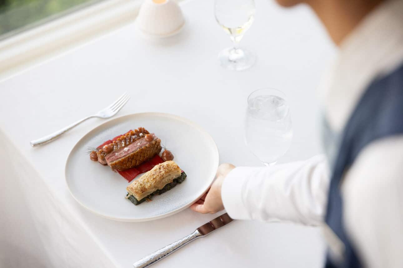 A person in formal attire is seated at a white table with a gourmet meal on a white plate, next to a fork, a glass of water, and a wine glass—evoking the refined dining experience of twenty-eight Atlantic.
