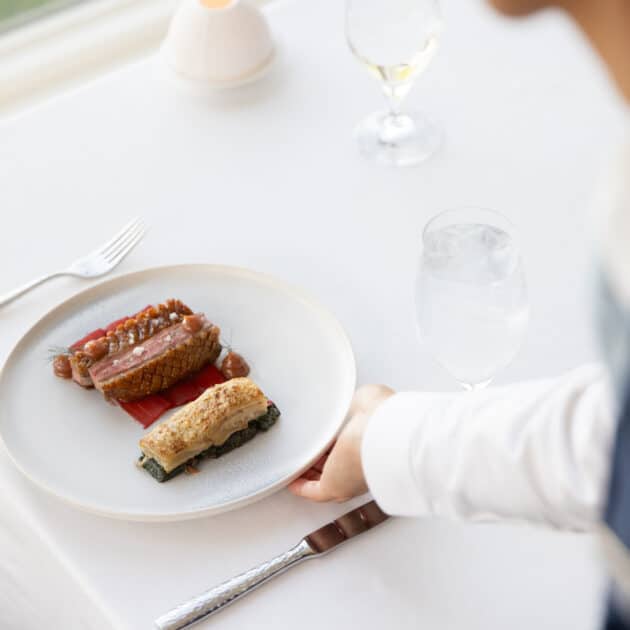 A person in formal attire is seated at a white table with a gourmet meal on a white plate, next to a fork, a glass of water, and a wine glass—evoking the refined dining experience of twenty-eight Atlantic.