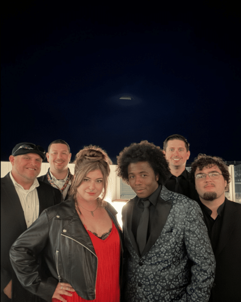 Six people dressed in formal and semi-formal attire pose together outdoors at night during a Cape Cod Arts Festival, with a dark sky and faint background lights behind them.