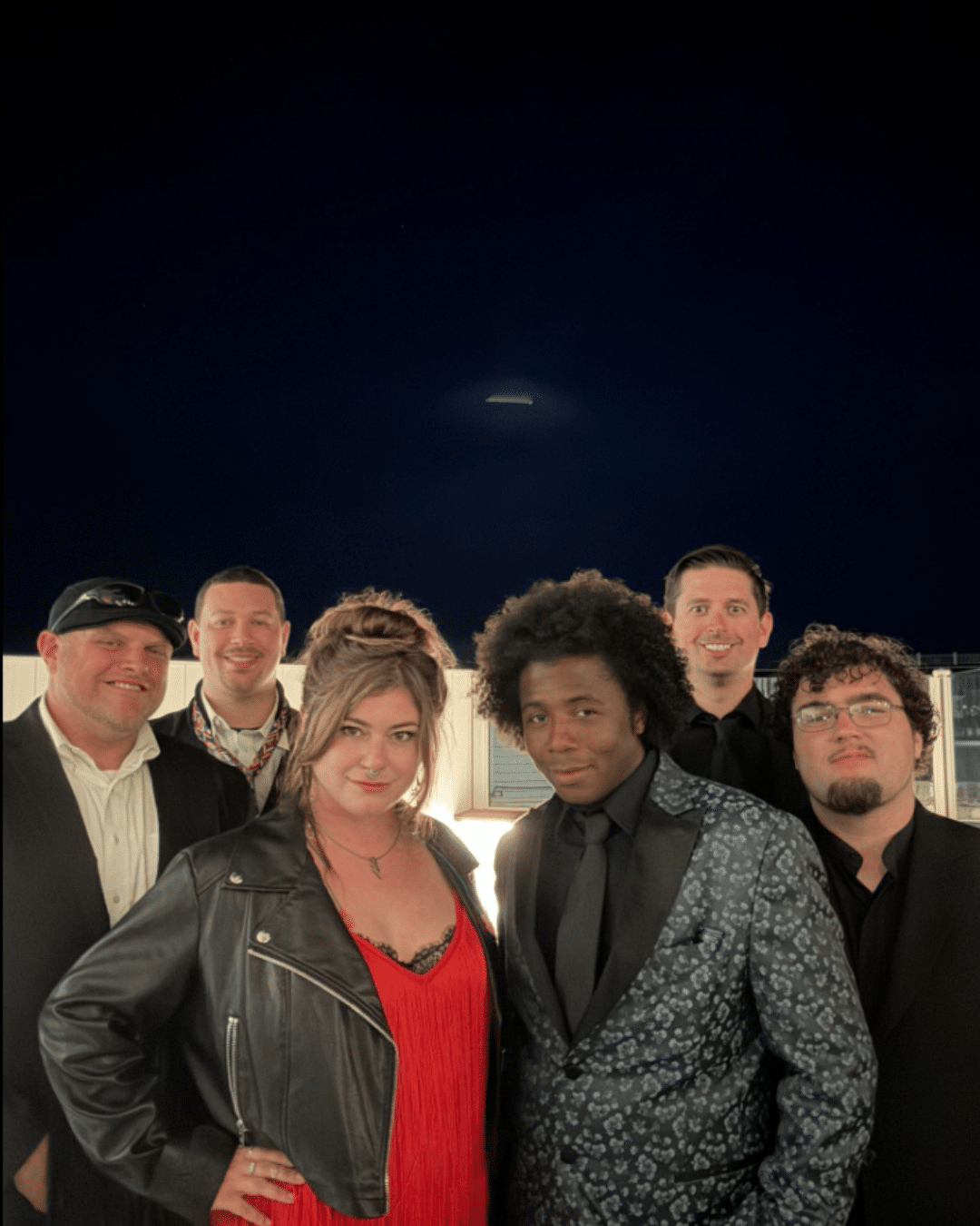 Six people dressed in formal and semi-formal attire pose together outdoors at night during a Cape Cod Arts Festival, with a dark sky and faint background lights behind them.