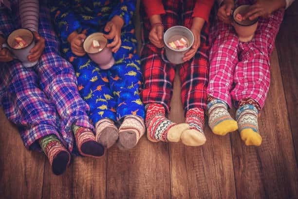 Four children in colorful pajamas and patterned socks sit on a wooden floor, holding mugs of hot chocolate with marshmallows, excitedly chatting about the upcoming Merry Market and fireworks at the annual event.