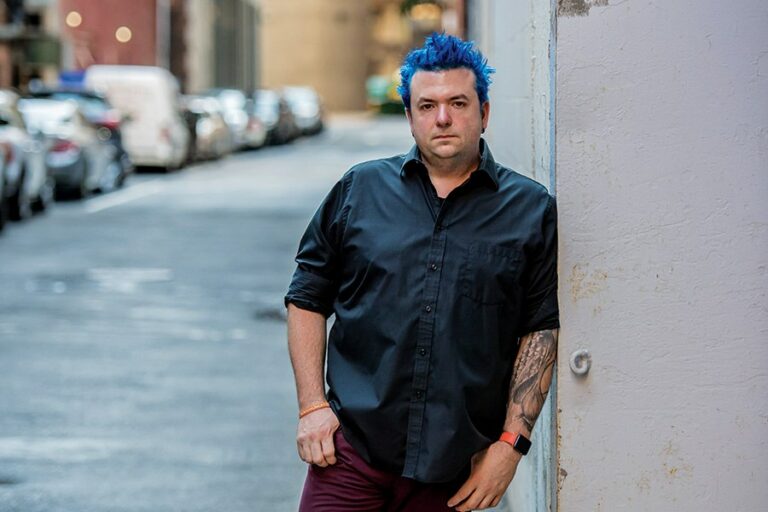 Man with blue hair and black shirt posing against a building.
