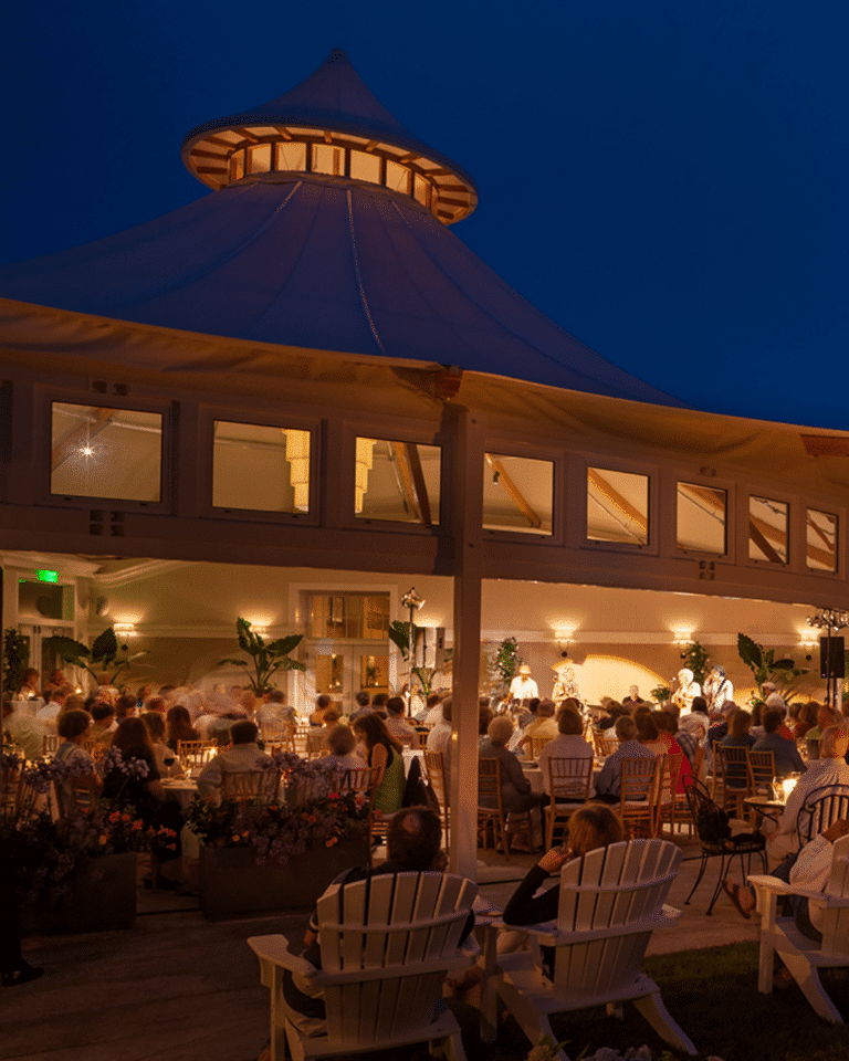 A crowd sits in white chairs outside a pavilion with a tent-like roof at night, enjoying a Jazz Festival performance under warm lighting on Cape Cod.