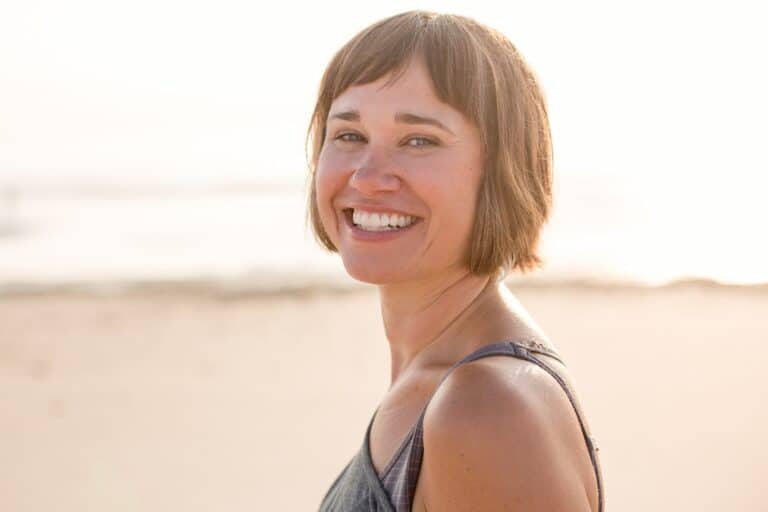 A woman with short brown hair smiles while standing on a beach, with soft sunlight and sand in the background.