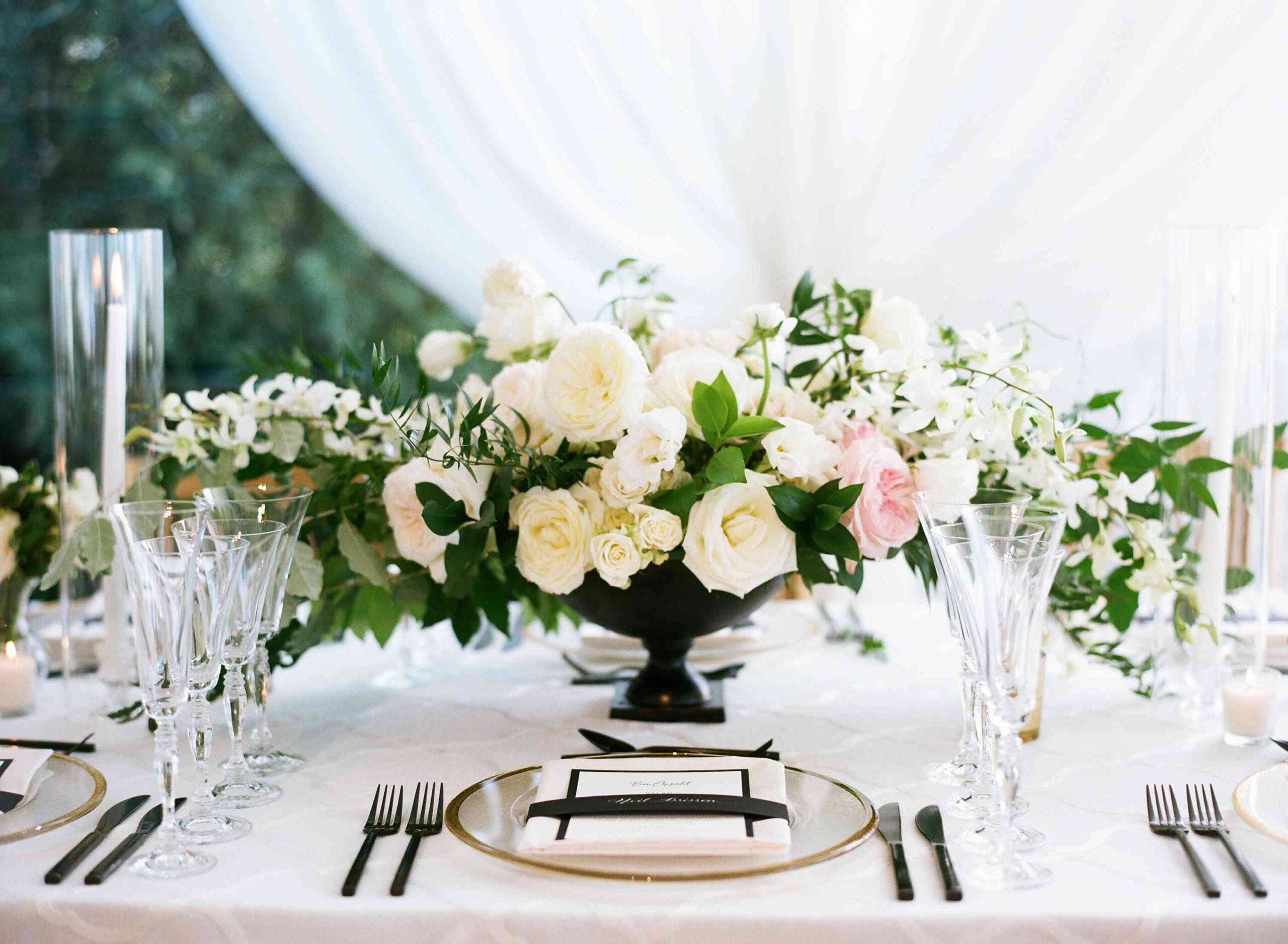 Elegant table setting with crystal glassware, black cutlery, and a centerpiece of white and pale pink flowers in a black vase on a white tablecloth—perfect for a refined dining experience at a restaurant Cape Cod.