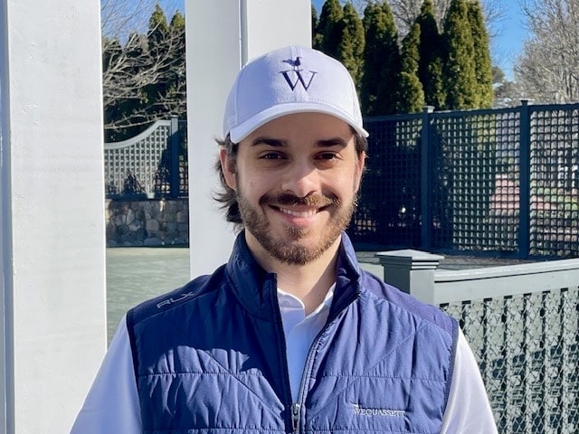 A man with a beard and mustache wearing a white cap, white shirt, and blue vest stands outside near a white pillar and trellis on a sunny day, looking ready for some racquet sports action.