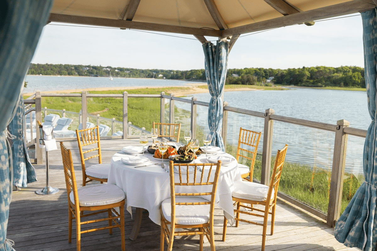 A round table with a white tablecloth and six chairs is set for a meal under a private cabana gazebo on a deck overlooking a river and green landscape, creating an inviting atmosphere perfect for an intimate clambake.