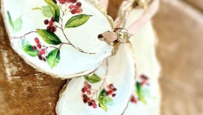 Three decorative oyster shells, inspired by Holiday Baking Classic motifs, feature painted red berries and green leaves, edged in gold. They lie on a wooden surface with pink ribbon loops attached.