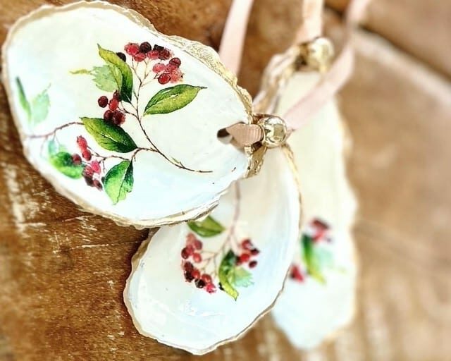 Three decorative oyster shells, inspired by Holiday Baking Classic motifs, feature painted red berries and green leaves, edged in gold. They lie on a wooden surface with pink ribbon loops attached.