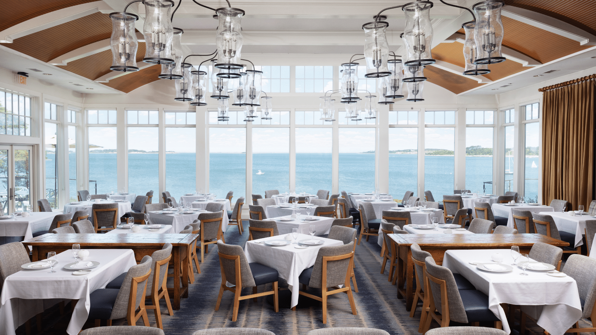 A bright restaurant dining room with tables set for service, large windows offer a view of water and distant land, and modern glass light fixtures hang from the ceiling.