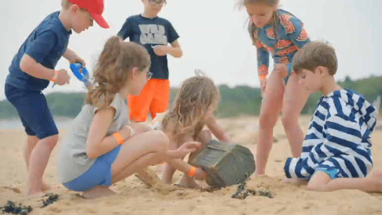 Six children gather around a bucket on the sandy Outer Beach, examining its contents with interest. Trees and water are visible in the background, highlighting the group’s beach journey of discovery.