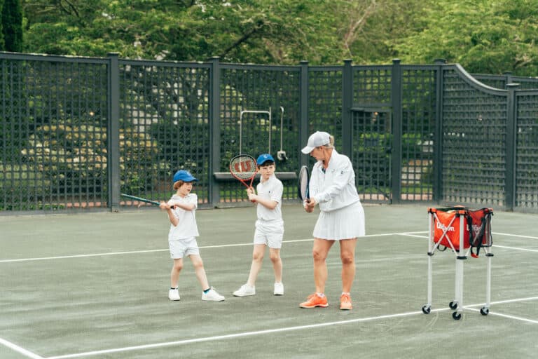 A tennis coach instructs two young children holding rackets on a tennis court, introducing them to the fundamentals of racquet sports. A basket of tennis balls stands nearby.