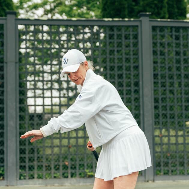 A person in a white tennis outfit and cap prepares to hit a tennis ball on an outdoor court with a lattice fence in the background, showcasing their skills in racquet sports.