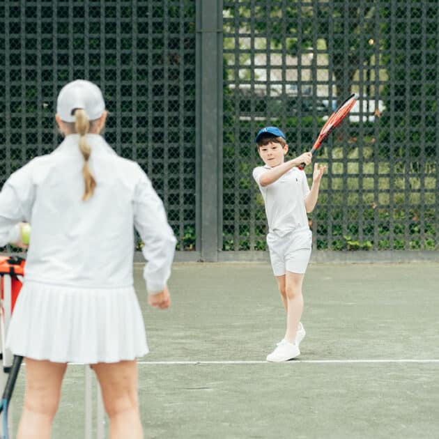 A young boy in sportswear swings a tennis racket, hitting a ball, while an adult in white tennis attire observes on an outdoor court, enjoying the excitement of racquet sports.
