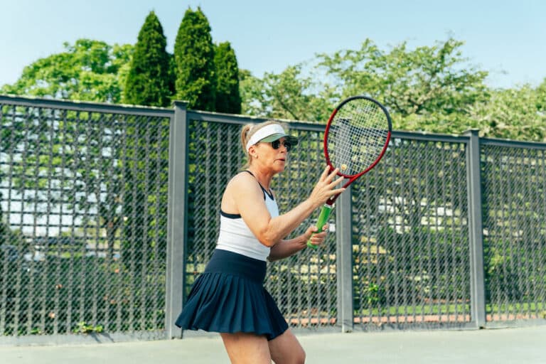 A woman in athletic attire holds a tennis racket, preparing to hit a ball on an outdoor tennis court surrounded by greenery—a perfect scene for racquet sports enthusiasts.
