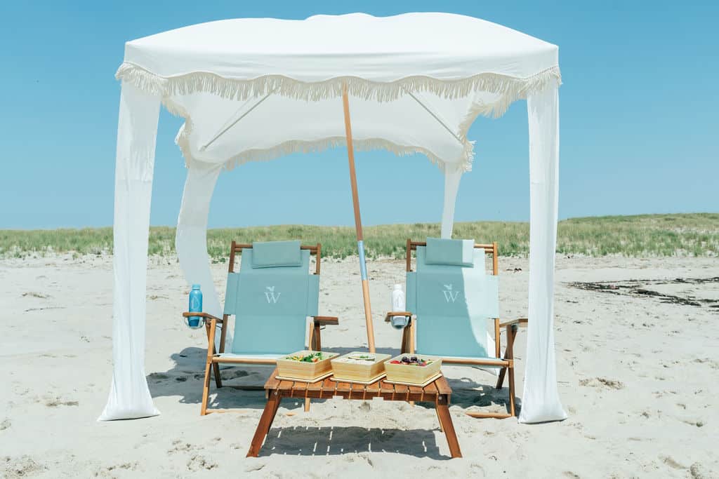 Two wooden beach chairs under a white canopy on sandy beach, with a table holding snacks and drinks, set against a grassy dune and blue sky—a perfect scene for your Outer Beach travel adventure.