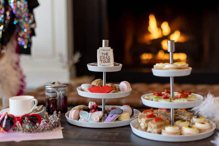 Three-tiered trays display assorted pastries, cookies, and sandwiches for Afternoon SwifTea in front of a lit fireplace. A cup, teapot, sunglasses, and accessories are arranged on the table for a cozy afternoon tea experience.