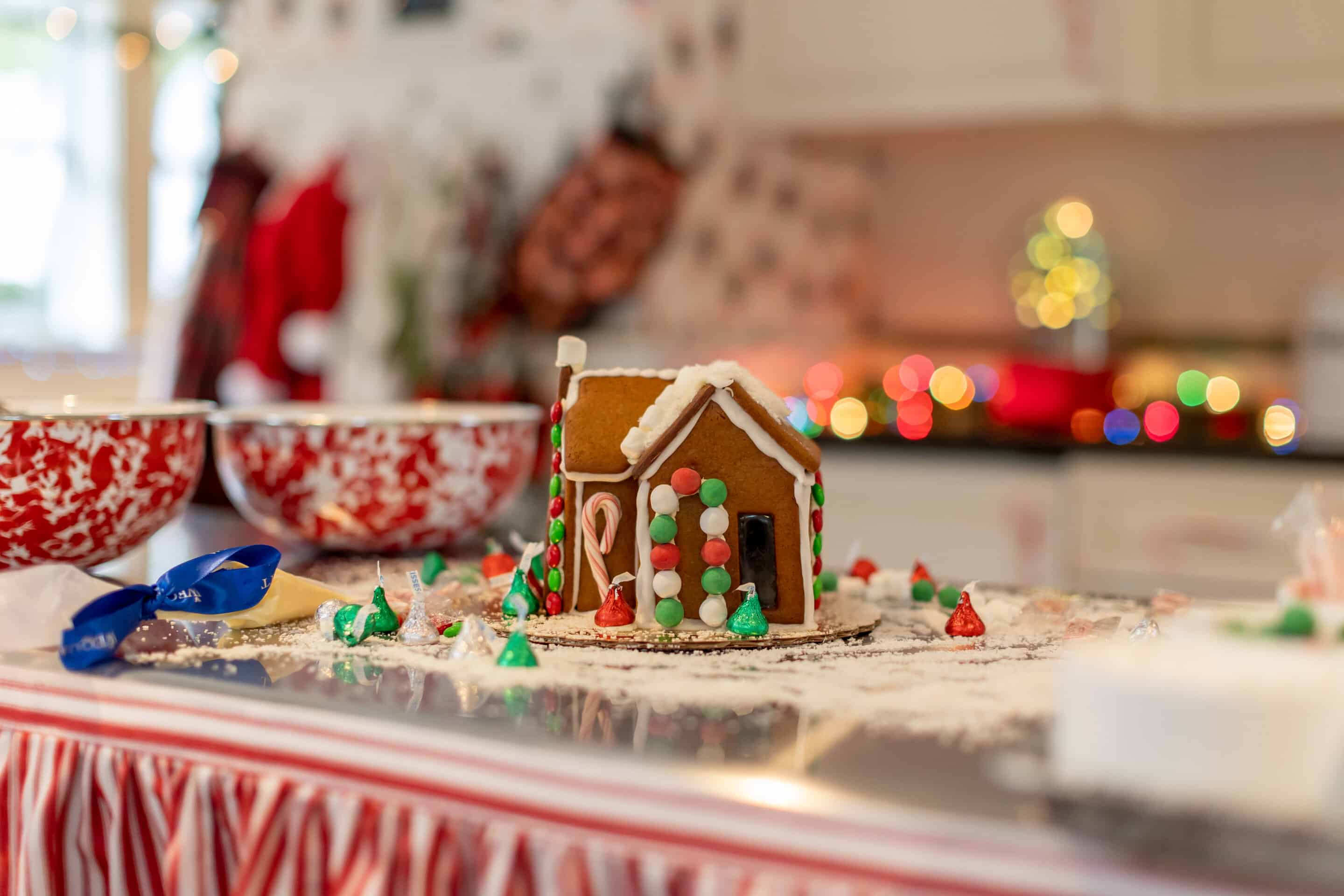 A decorated gingerbread house sits on a kitchen counter surrounded by icing, candy, and mixing bowls, with holiday lights blurred in the background.